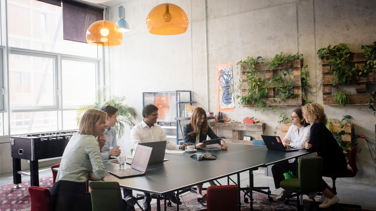 A group of people in a modern meeting room with laptops, plants, and colorful pendant lights.