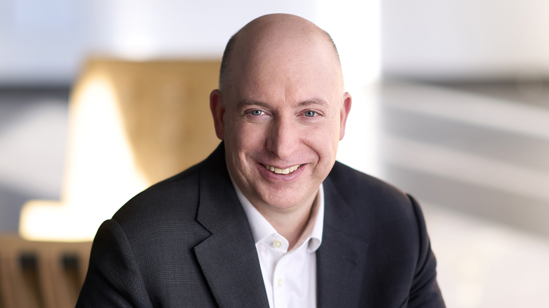 Ein Mann mit Glatze trägt einen dunklen Anzug und sitzt in einem hellen Raum mit verschwommenem Hintergrund.