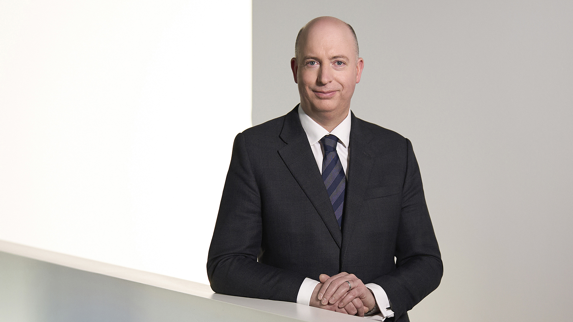 Ein Mann in einem Anzug steht an einer weißen Wand und hält seine Hände auf einer Tischkante.