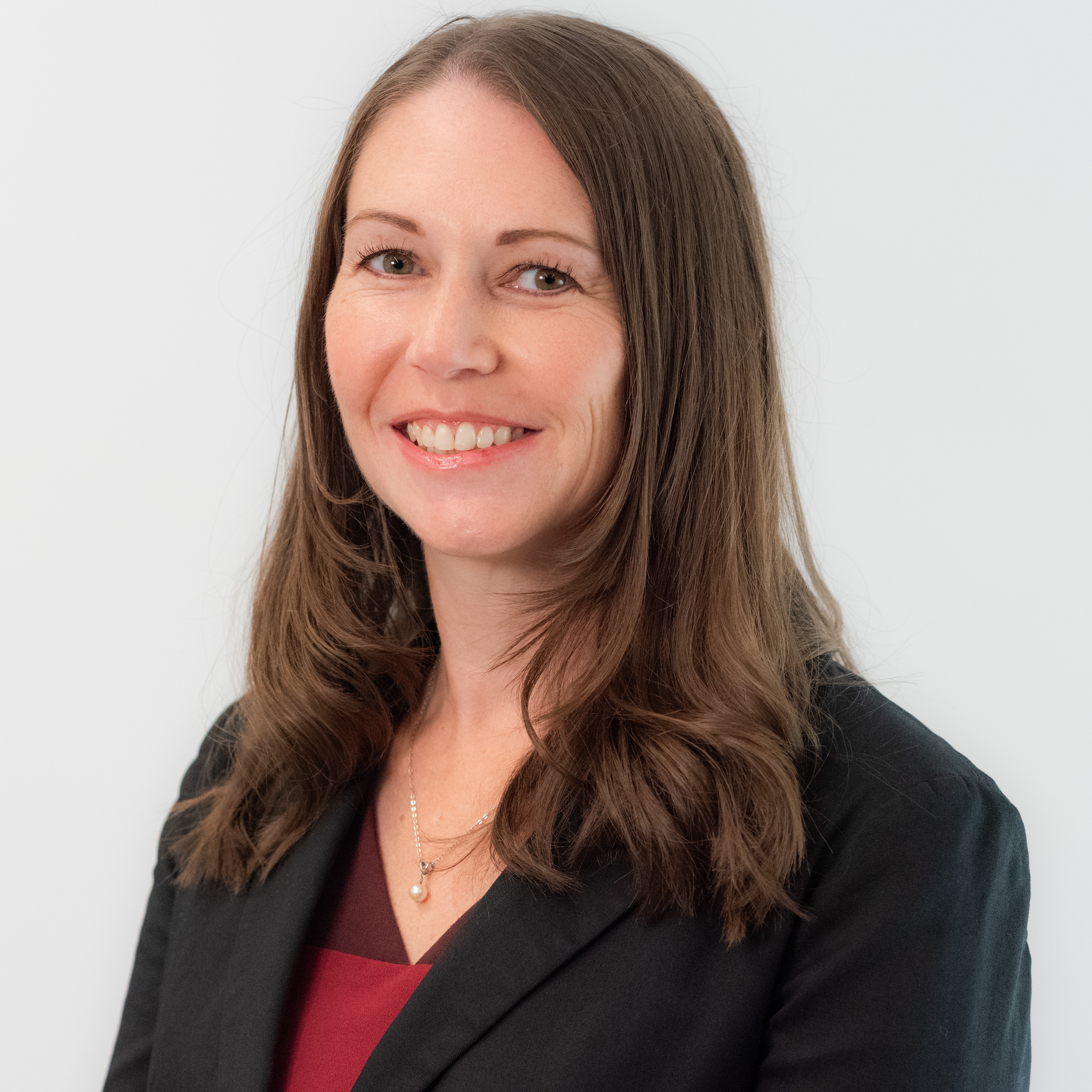 A woman with long brown hair wearing a black blazer and a maroon top, set against a plain white background.