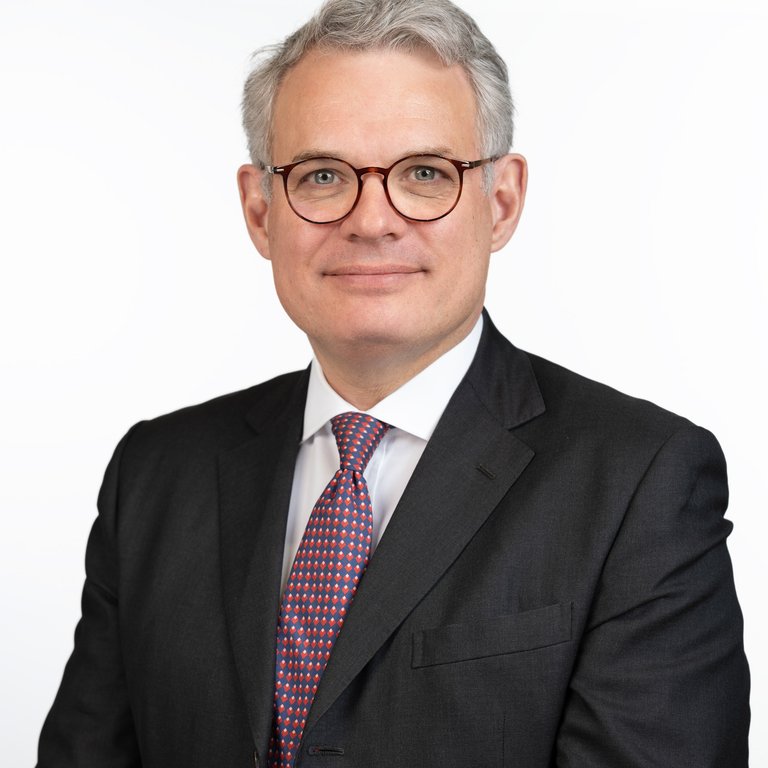 A man in a dark suit and patterned tie stands against a white background, face obscured.
