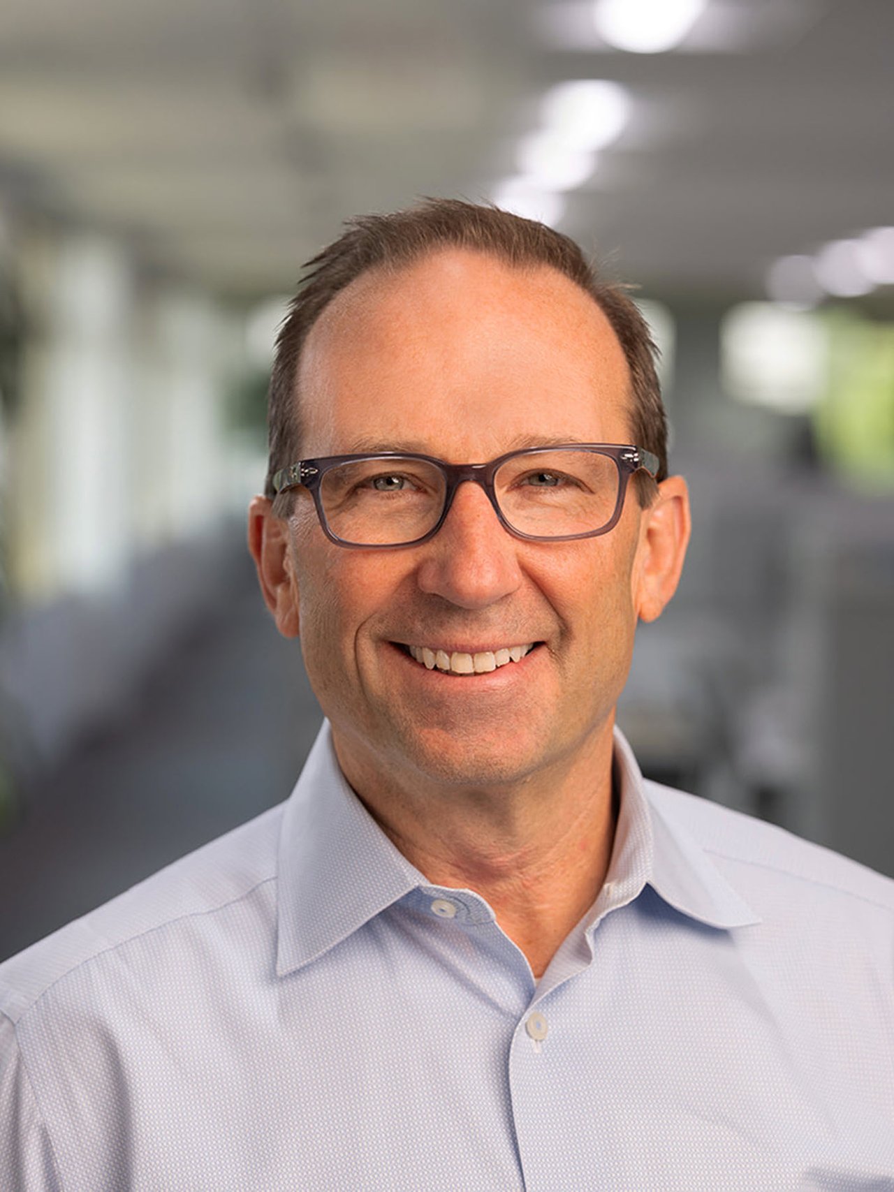 A smiling man in a light blue shirt stands in an office setting with blurred desks and plants in the background.