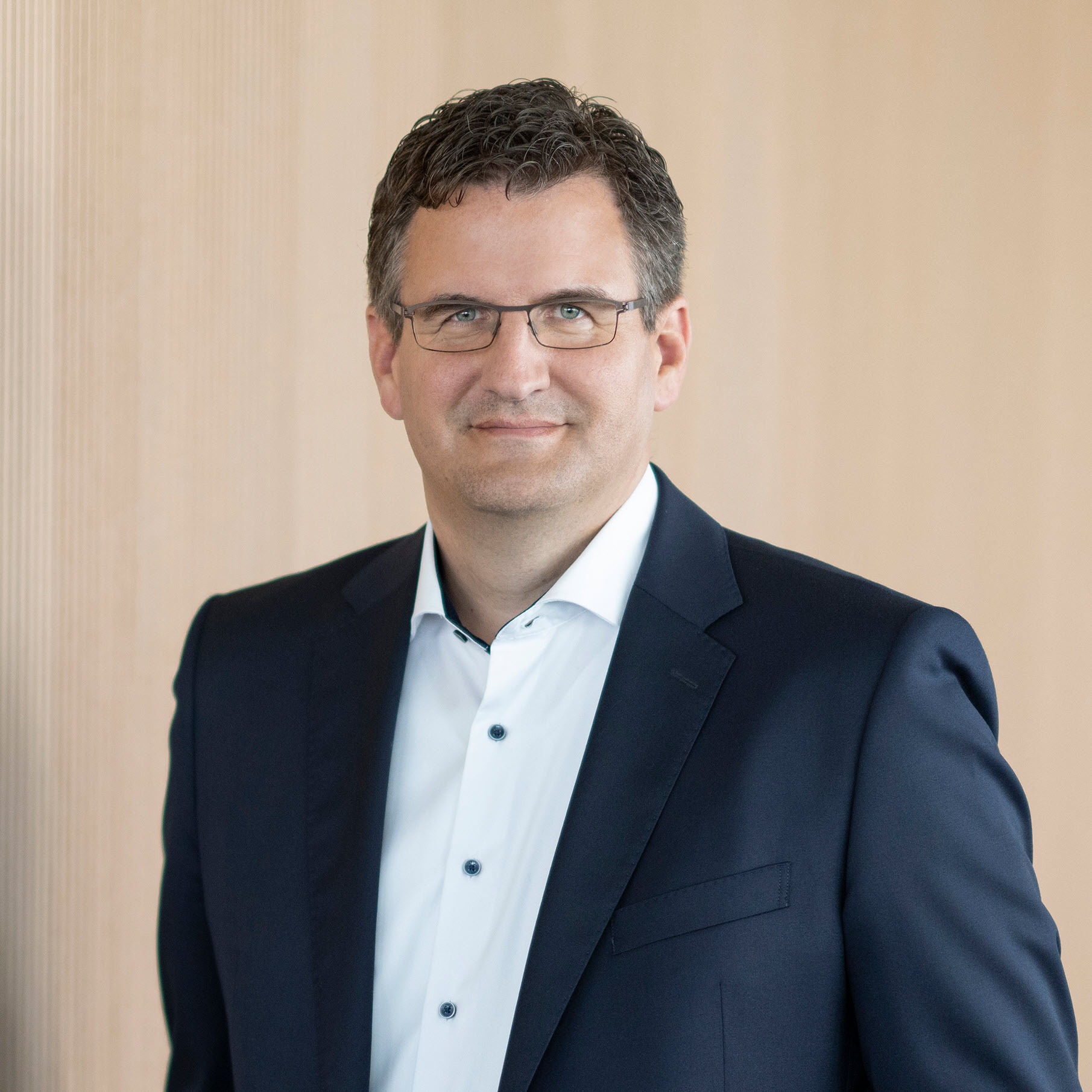 A man in a dark suit and white shirt stands against a light wooden background.
