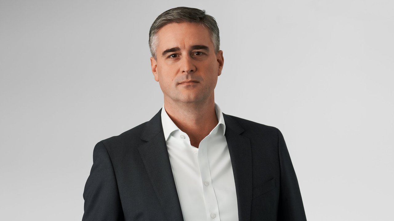 A man in a dark suit and white shirt stands against a plain gray background, looking directly at the camera.