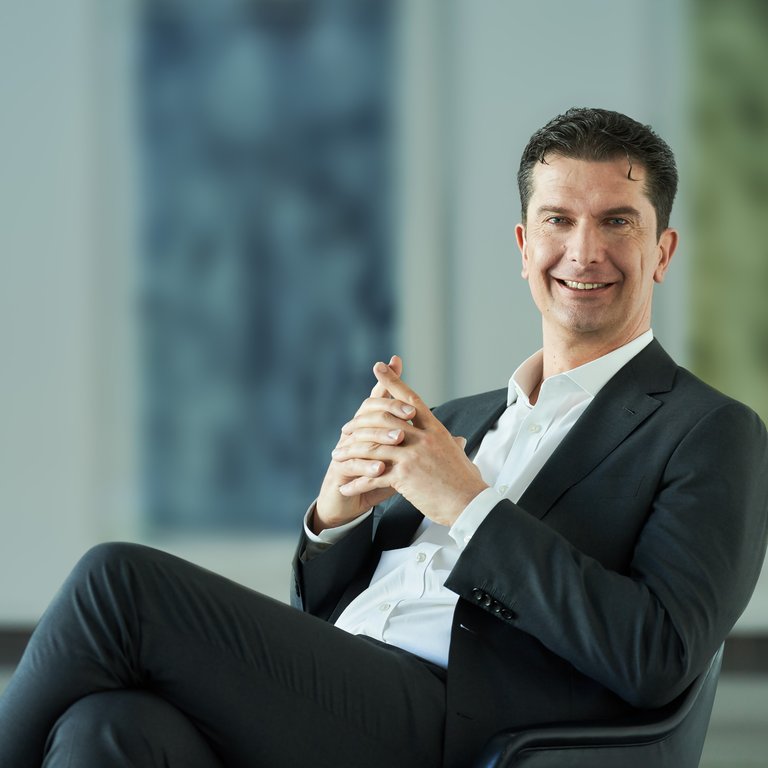 A person in a suit sits with hands clasped, in a modern interior with abstract art in the background.