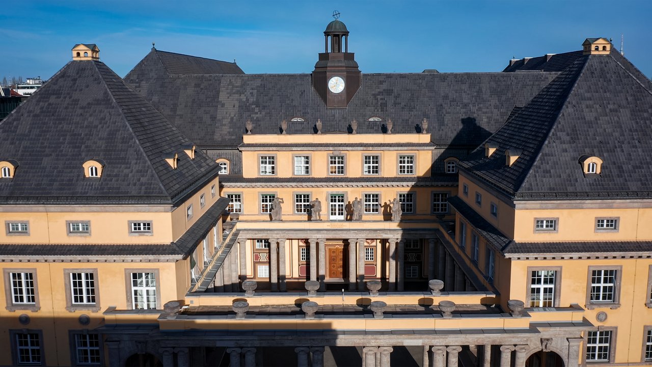 Aerial view of a large yellow building with a clock tower and multiple gabled roofs, featuring columns and statues.
