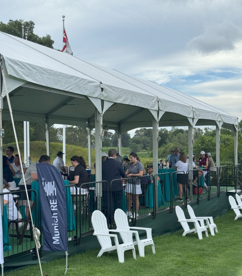 A tented area with people socializing, tables with teal covers, and white chairs on a grassy field.