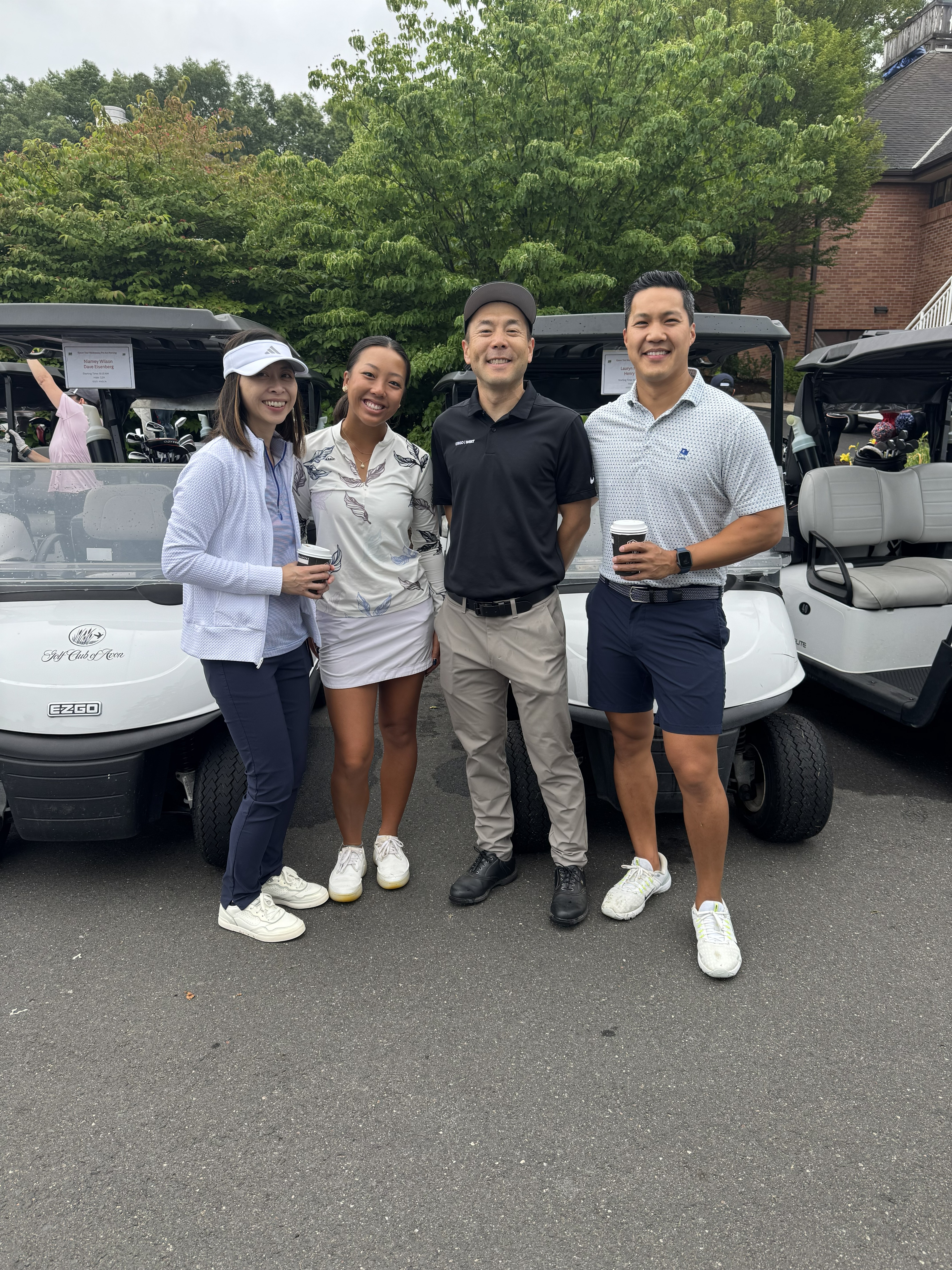 Four people pose together in front of golf carts, surrounded by greenery, holding coffee cups.