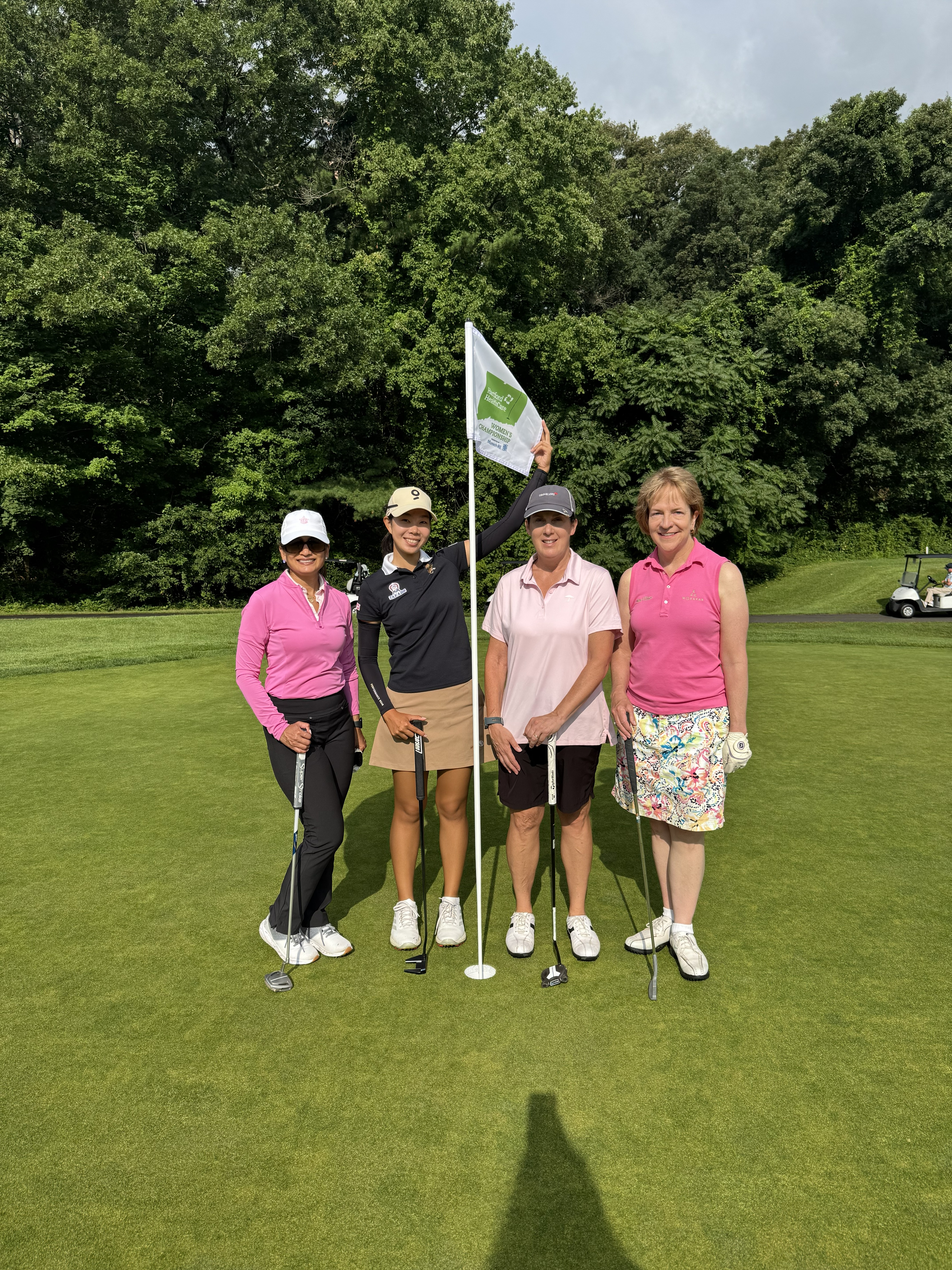 Four women stand on a golf course, smiling near a flagstick with a green background.