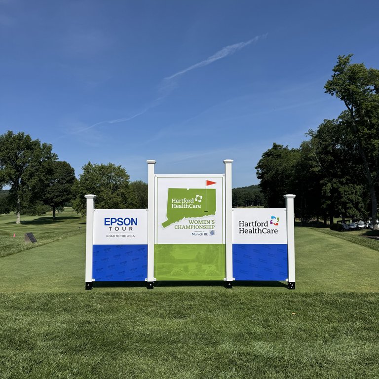 Signage for the Epson Tour and Hartford HealthCare Women's Championship on a golf course under a clear blue sky.