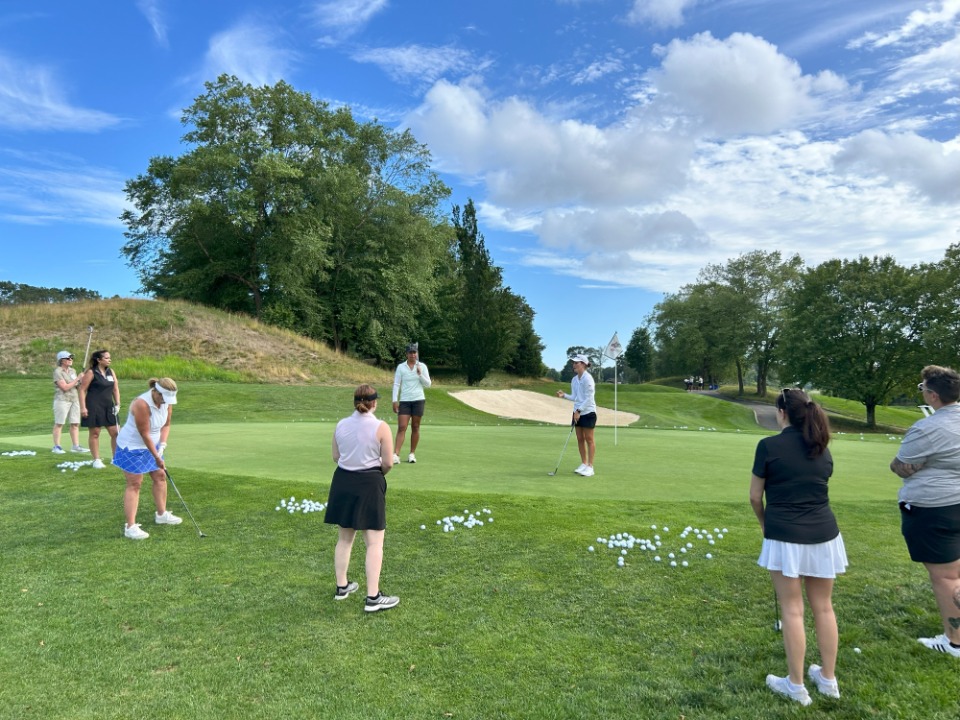 A group of women practicing golf on a green, with one instructor demonstrating a technique.