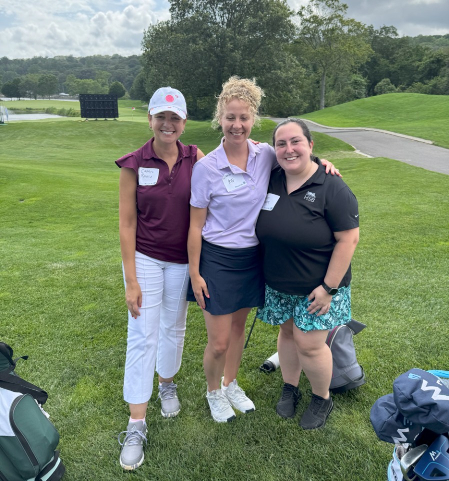 Three women stand together on a golf course, wearing golf attire and name tags, with green grass and trees in the background.