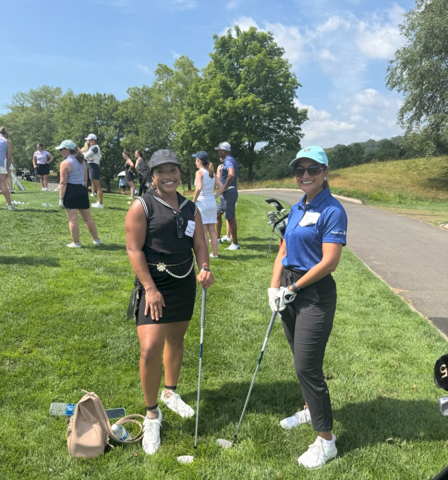 Two women stand on a golf course, holding clubs, with other golfers in the background under a clear blue sky.
