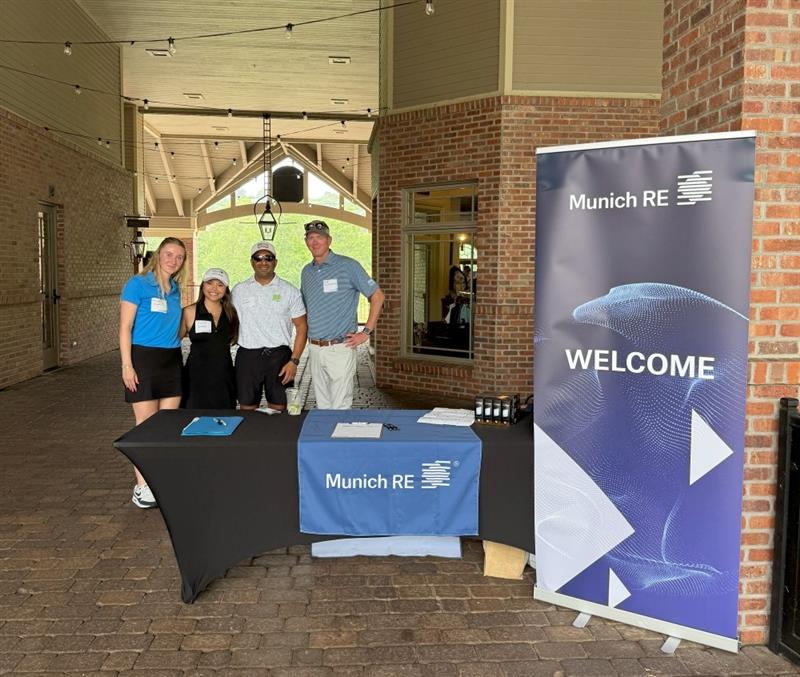 A group of four people stands by a welcome table with a Munich RE banner in a brick corridor.
