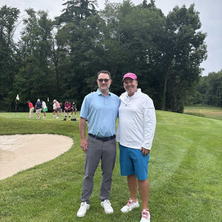 Two men stand on a golf course, smiling, with trees and other golfers in the background.