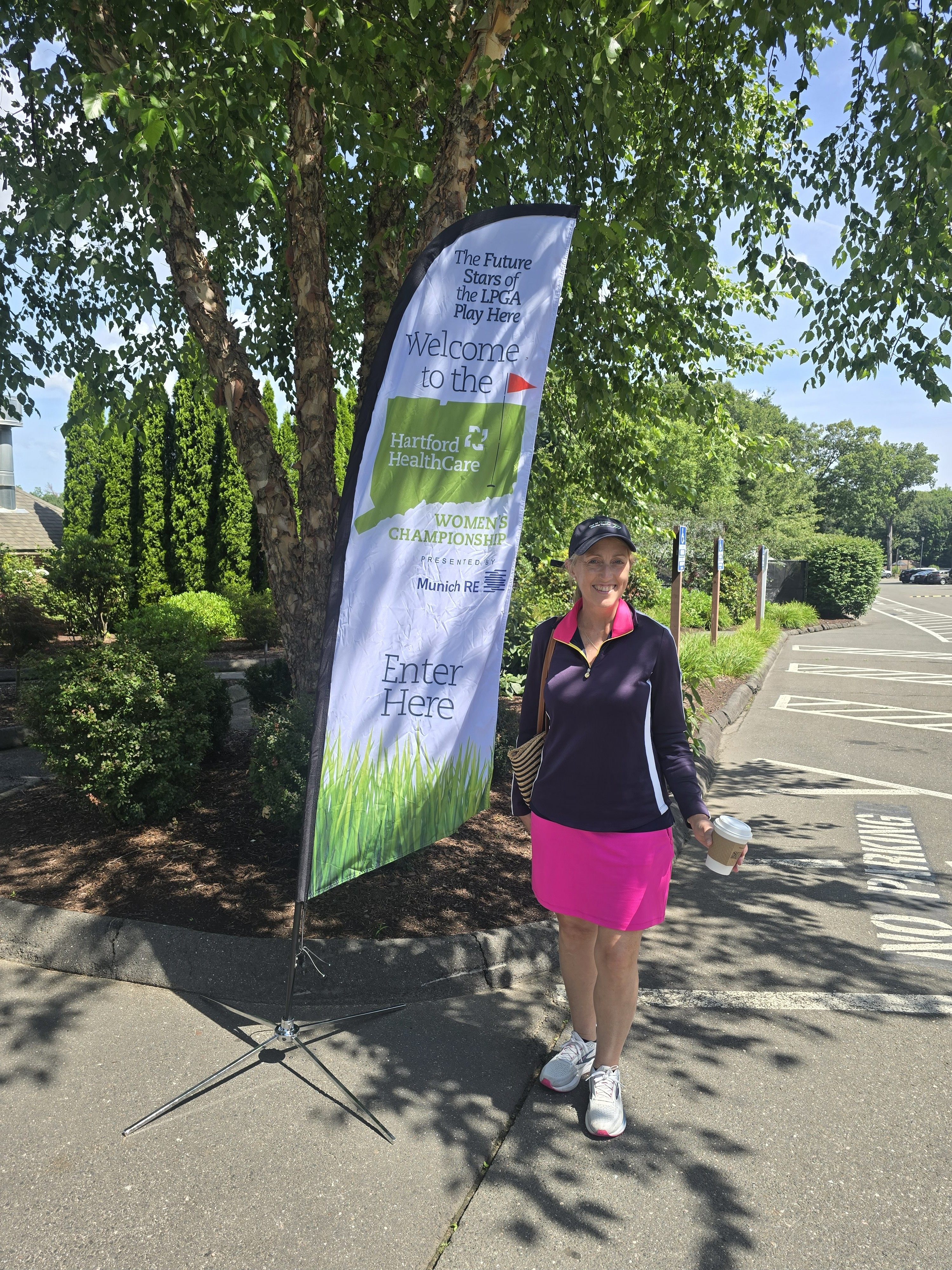 A woman in a pink skirt and dark top stands next to a banner for the Hartford HealthCare Women's Championship.