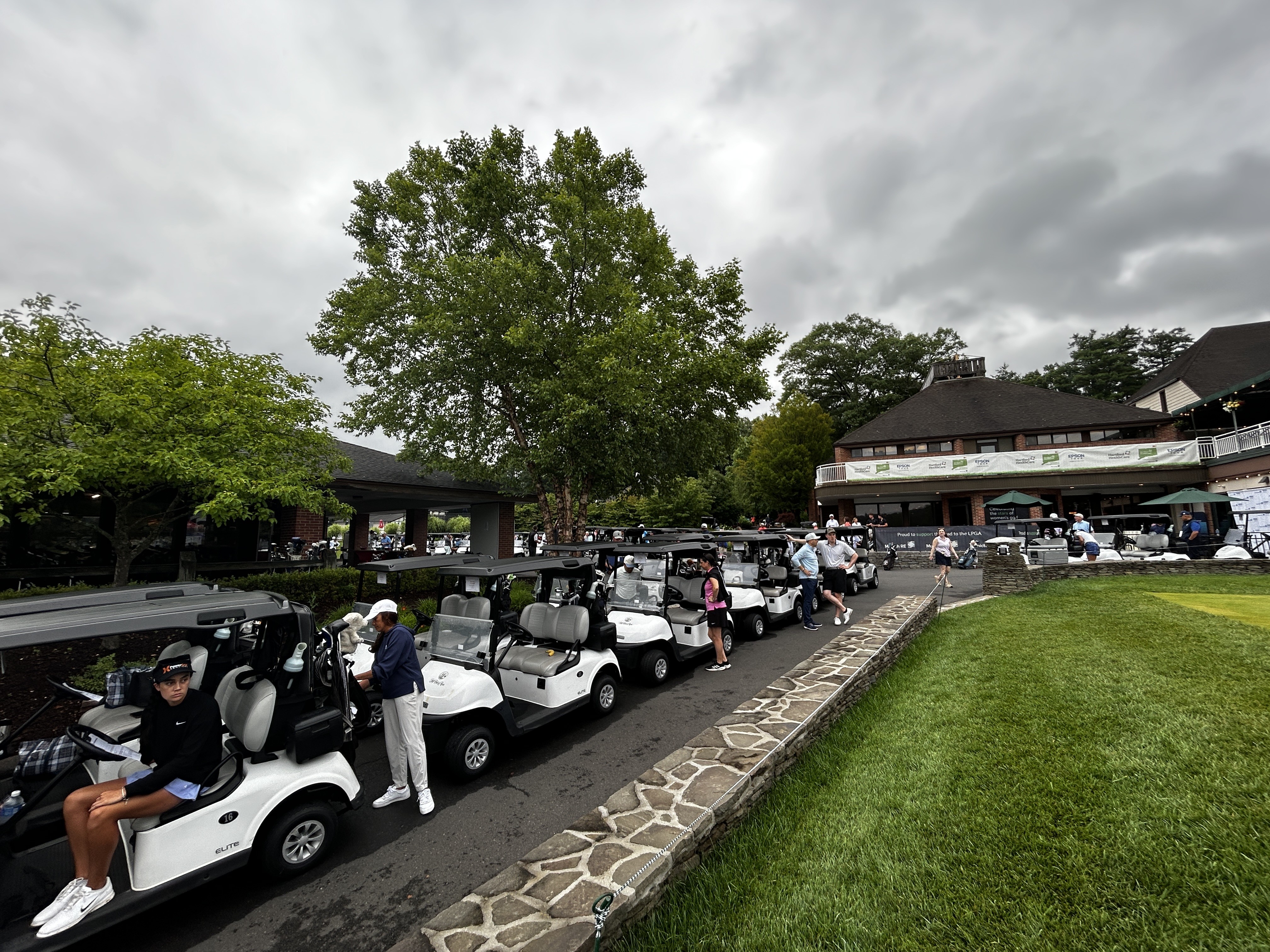 A line of golf carts parked near a clubhouse with people walking around under a cloudy sky.