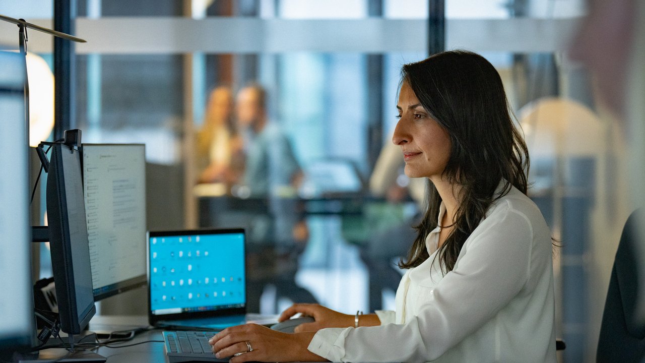 A woman works at a desk with two computer screens in a modern office setting.