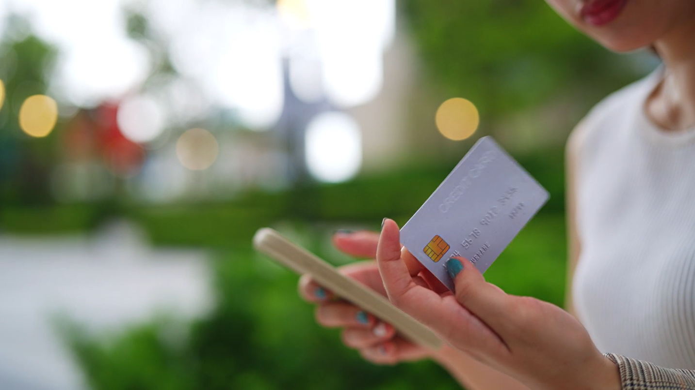A person holds a smartphone in one hand and a credit card in the other, with a blurred green background.