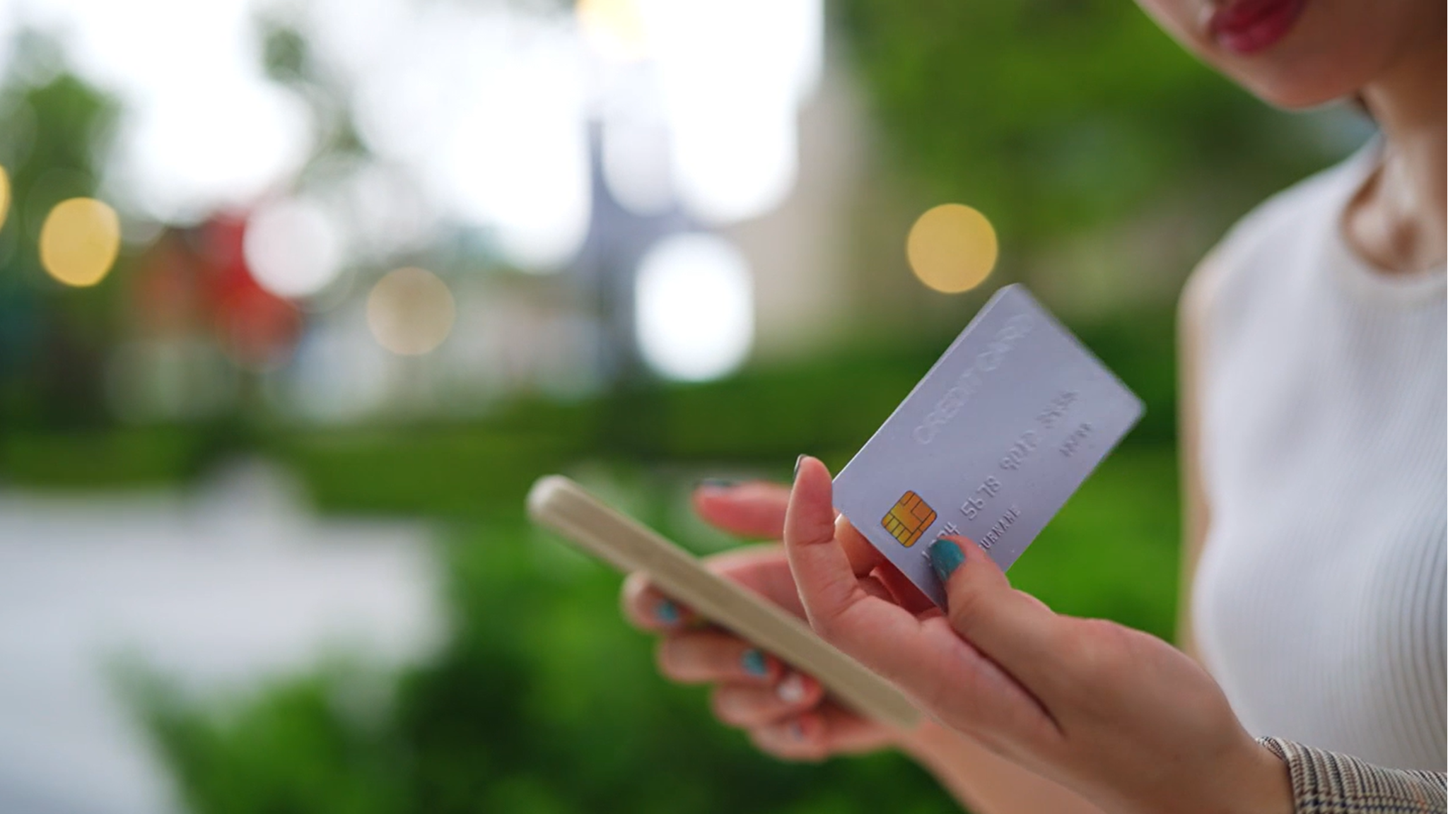 A person holds a smartphone in one hand and a credit card in the other, with greenery in the background.