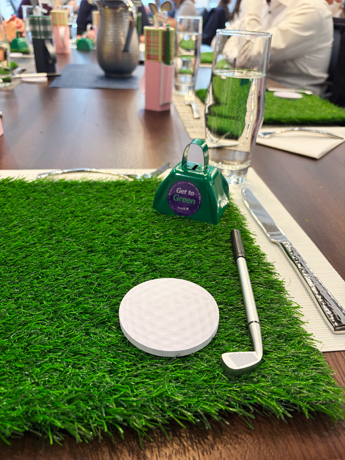 A golf-themed table setting with a golf ball, club, and a green grass mat, surrounded by glasses and decor.