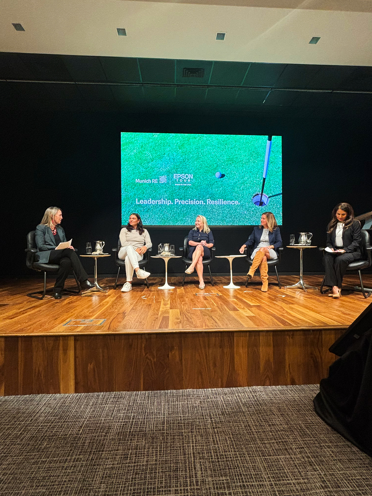 Four women sit on a stage discussing leadership, with a screen displaying "Leadership. Precision. Resilience." behind them.
