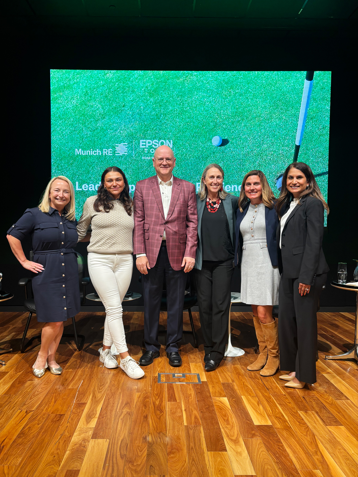 A group of six people posing together in front of a green backdrop with golf elements, on a wooden floor.