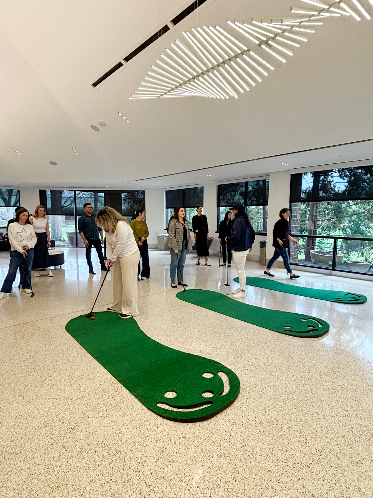 A group of people playing mini-golf indoors on green turf with smiling designs, under modern lighting.