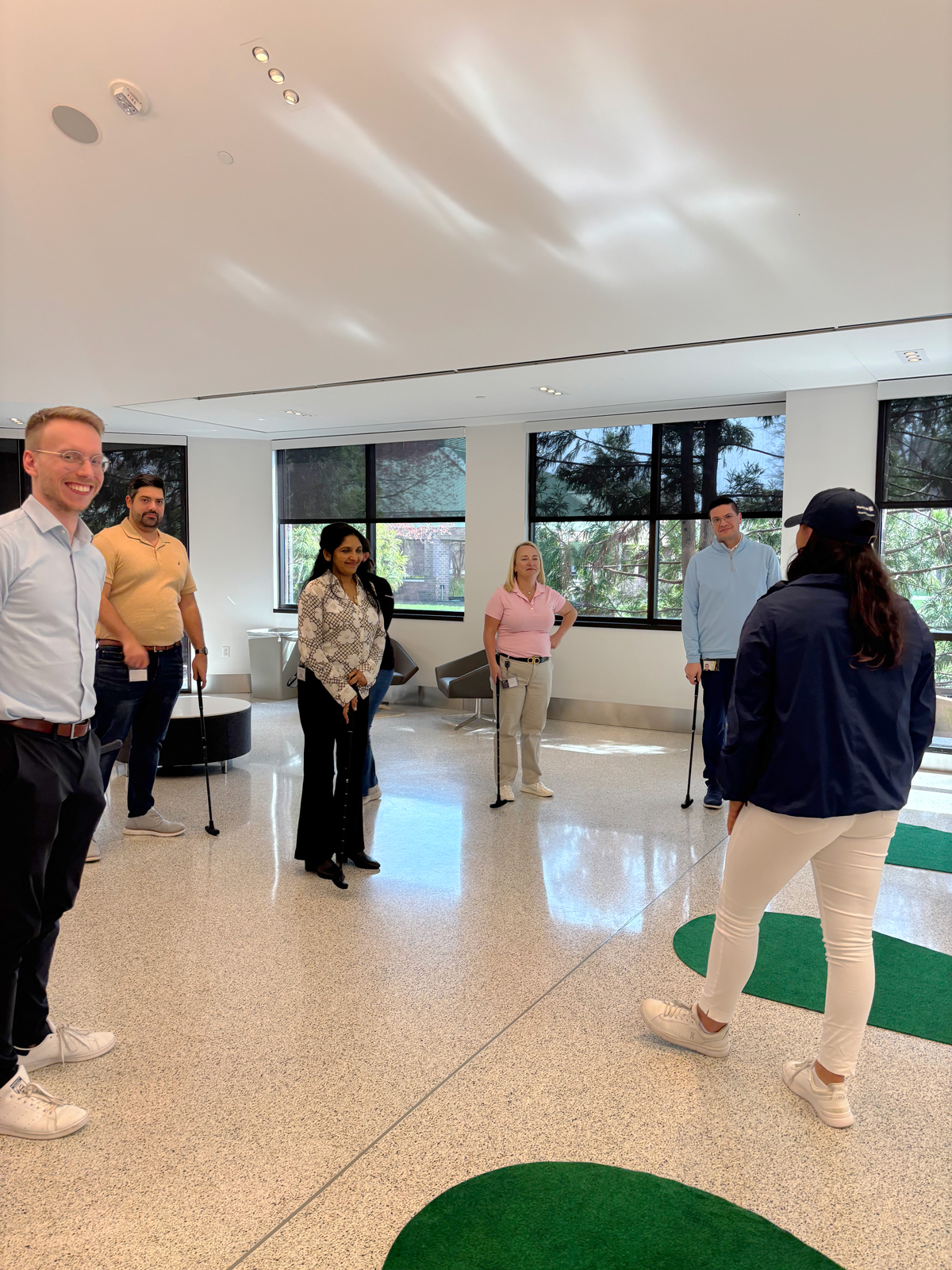 A group of six people standing indoors, some holding canes, in a well-lit room with green carpet patches.