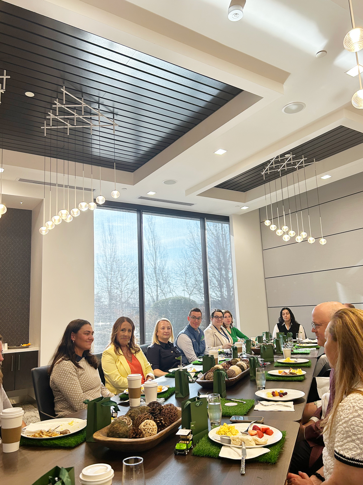A group of people seated at a long table with food, drinks, and decorative items in a well-lit meeting room.