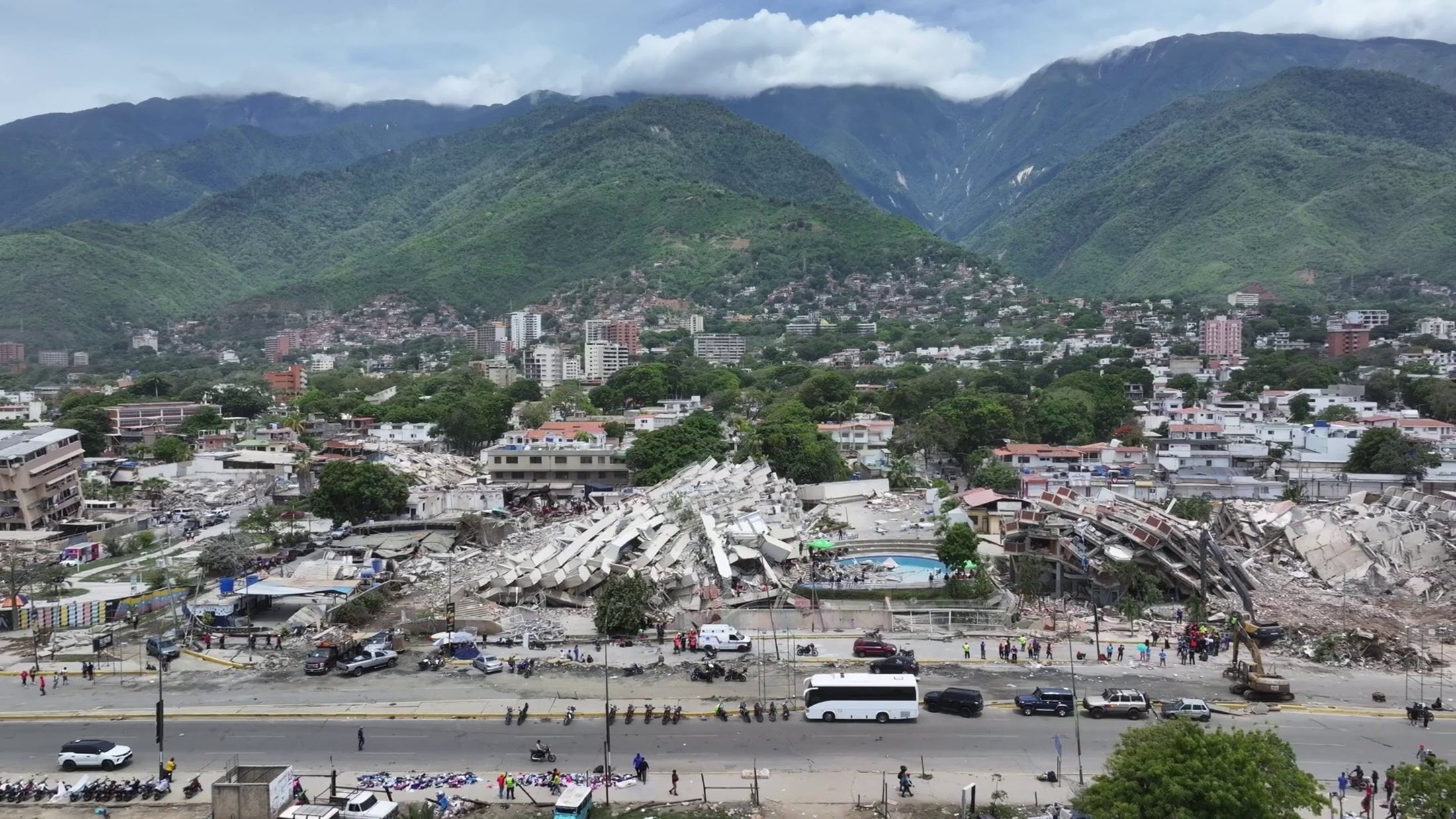 A view of a damaged city area with collapsed buildings, vehicles, and mountains in the background under a cloudy sky.