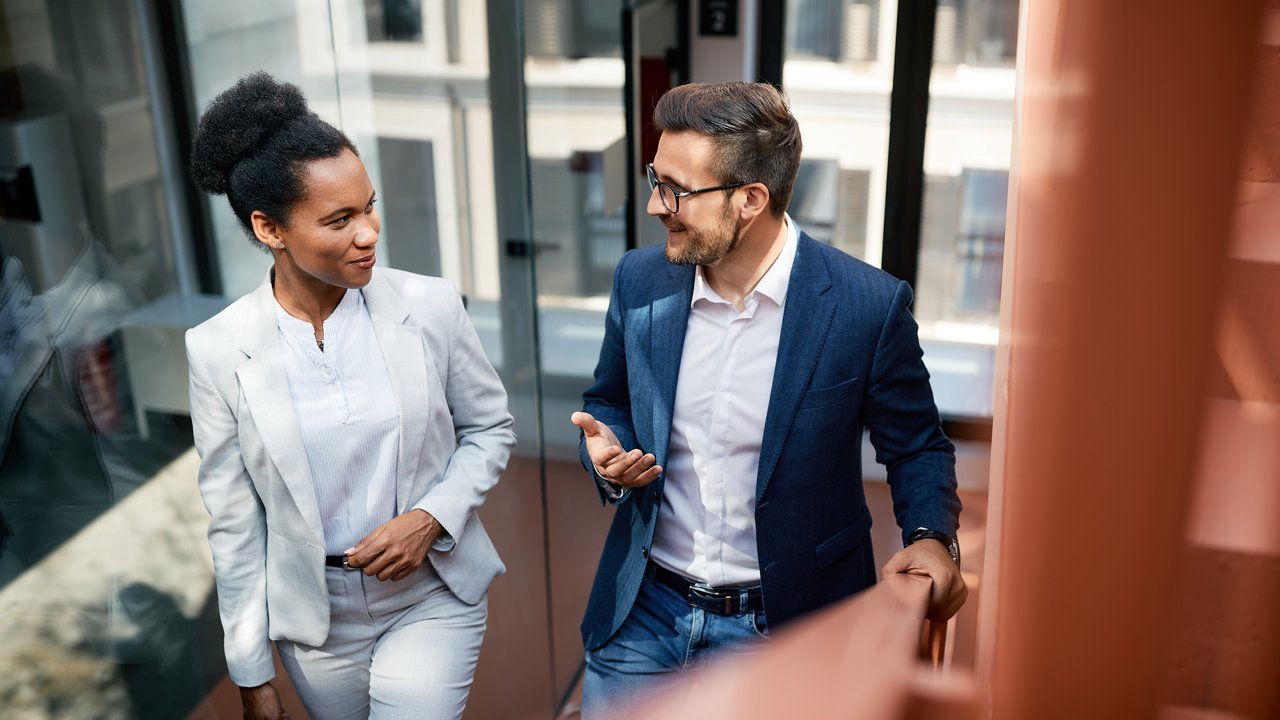 High angle view of a male and a female colleague are talking and smiling while walking up the stairs in a modern office building. High angle view of a male and a female colleague are talking and smiling while walking up the stairs in a modern office building.