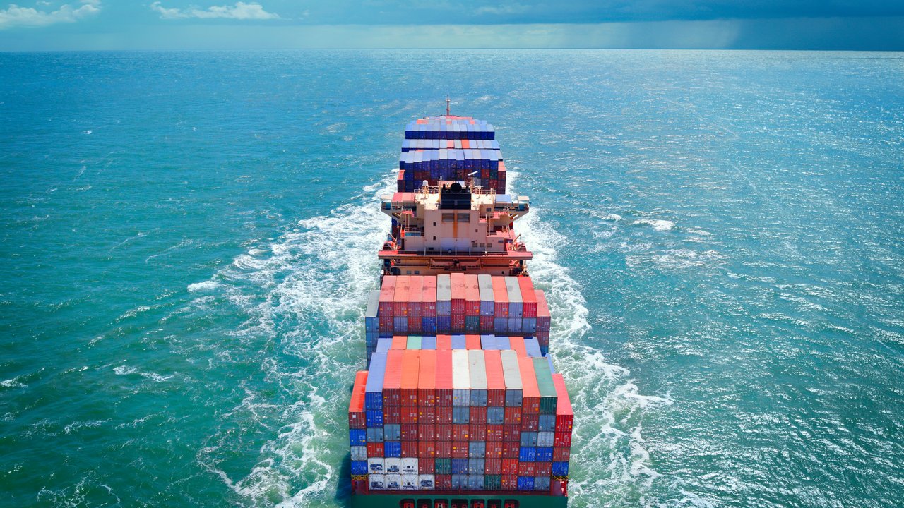 A large cargo ship loaded with colorful containers sailing on blue water under a partly cloudy sky.