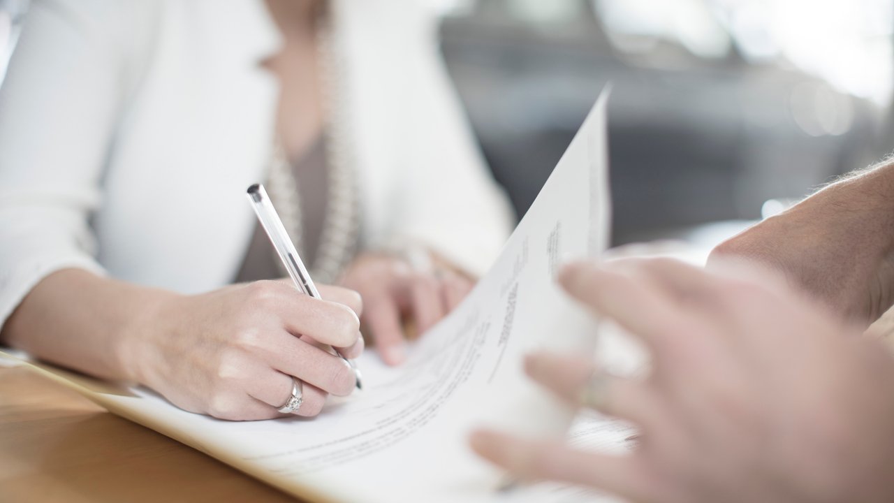 A person writes on a document while another hand holds a paper, with a blurred background of a car.