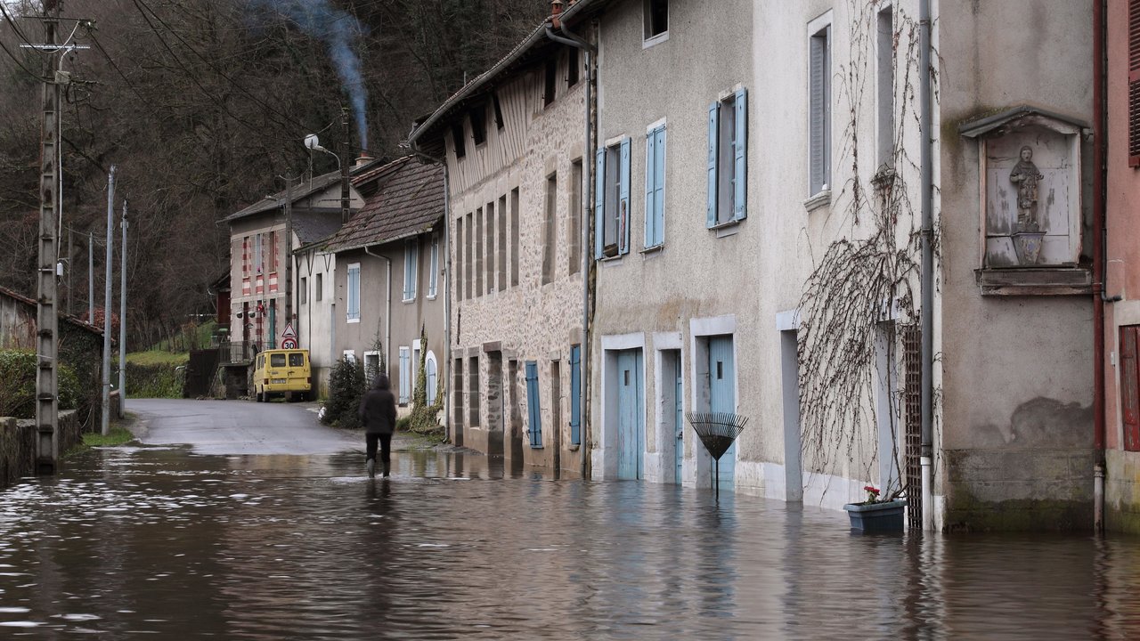 A flooded street with buildings, a person walking, and smoke rising from a chimney in the background.