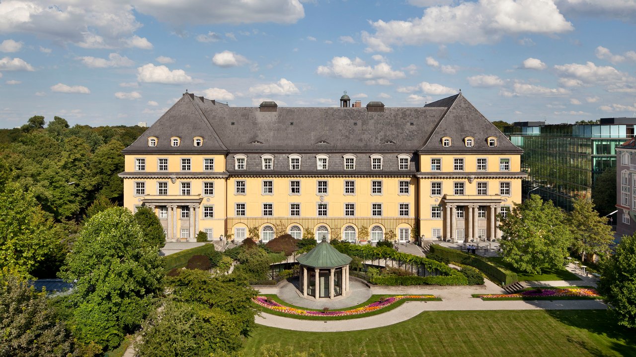 A large yellow building with a sloped roof, surrounded by greenery and a garden with a gazebo and flower beds.