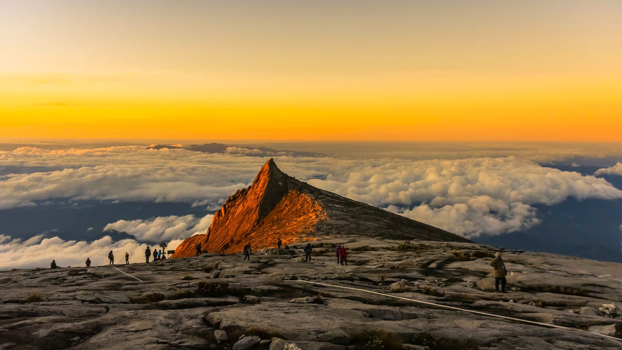 A mountain peak at sunset with hikers on rocky terrain above clouds, creating a scenic landscape.