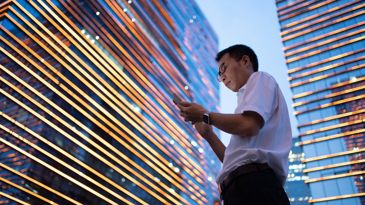 Planning portfolios with 1.5°C, 2°C, 3°C futures Businessman using smartphone in front of illuminated skyscrapers