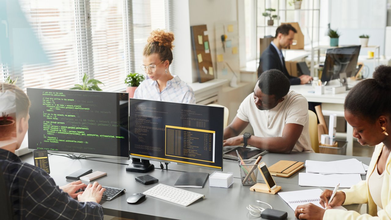 A modern office with four people working at desks, two using computers and two writing notes.