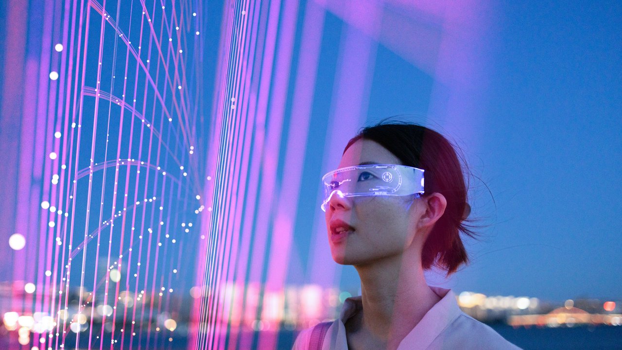 A woman wearing futuristic glasses stands in front of colorful light patterns against a twilight sky.