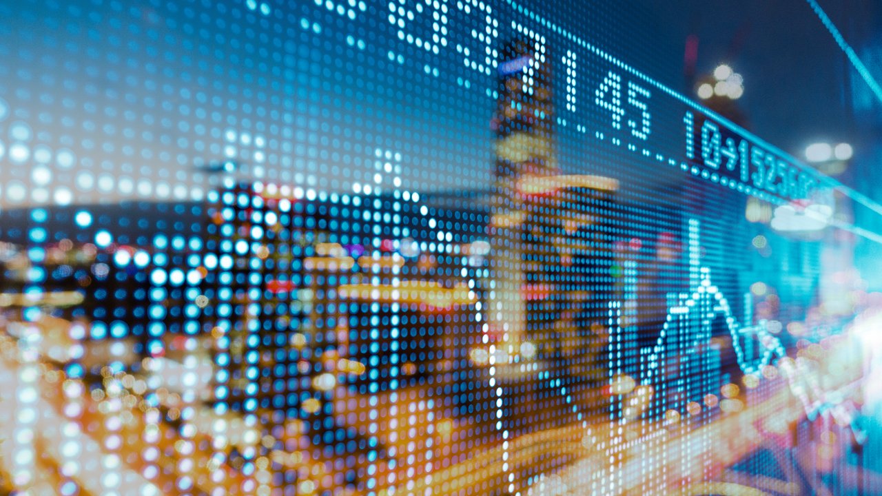 A digital display shows stock market data with graphs and numbers, blurred city lights in the background.
