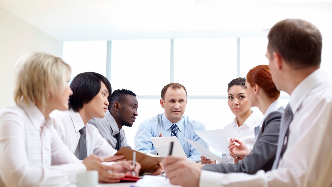 A group of six people in business attire engaged in a meeting around a table with papers and a coffee cup.