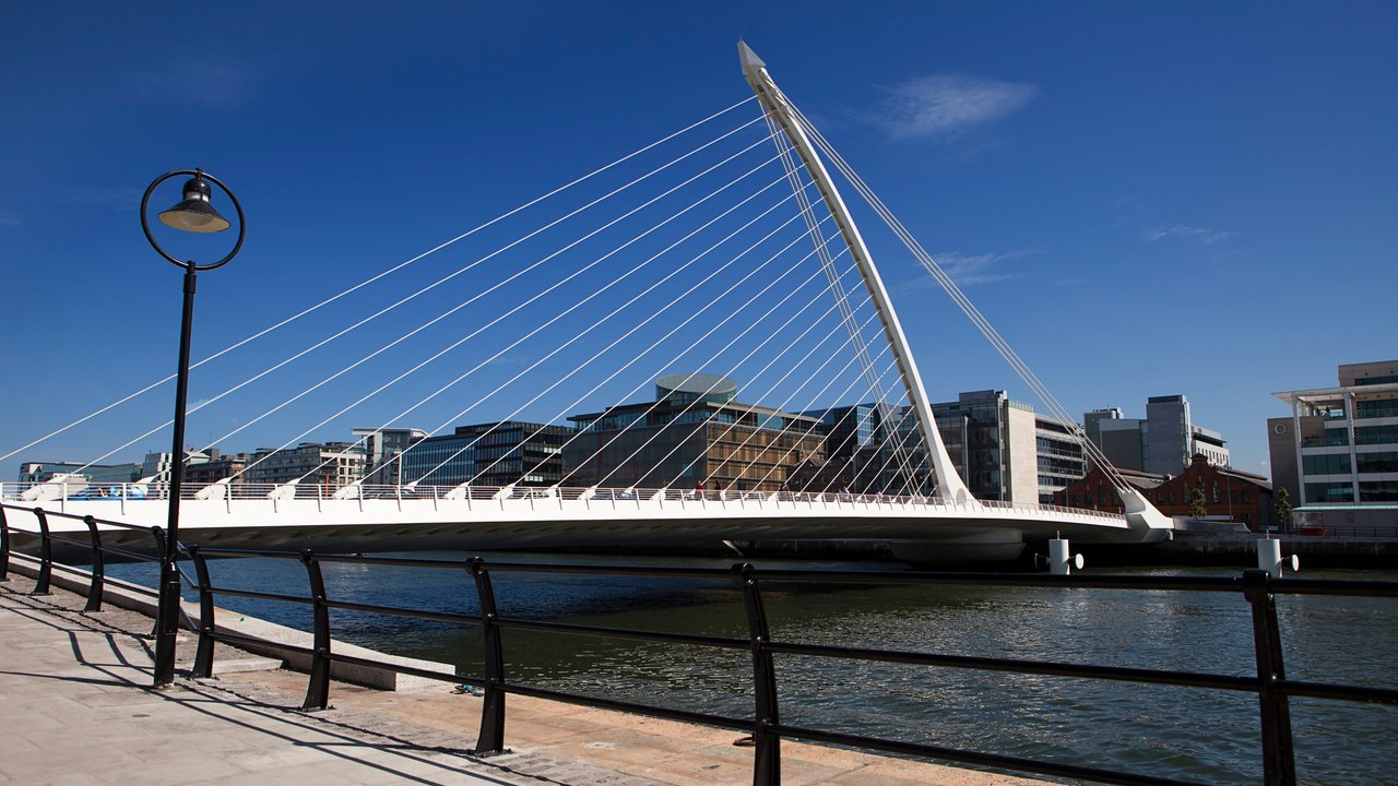 A modern cable-stayed bridge spans a river, with buildings and a lamp post visible along the walkway.