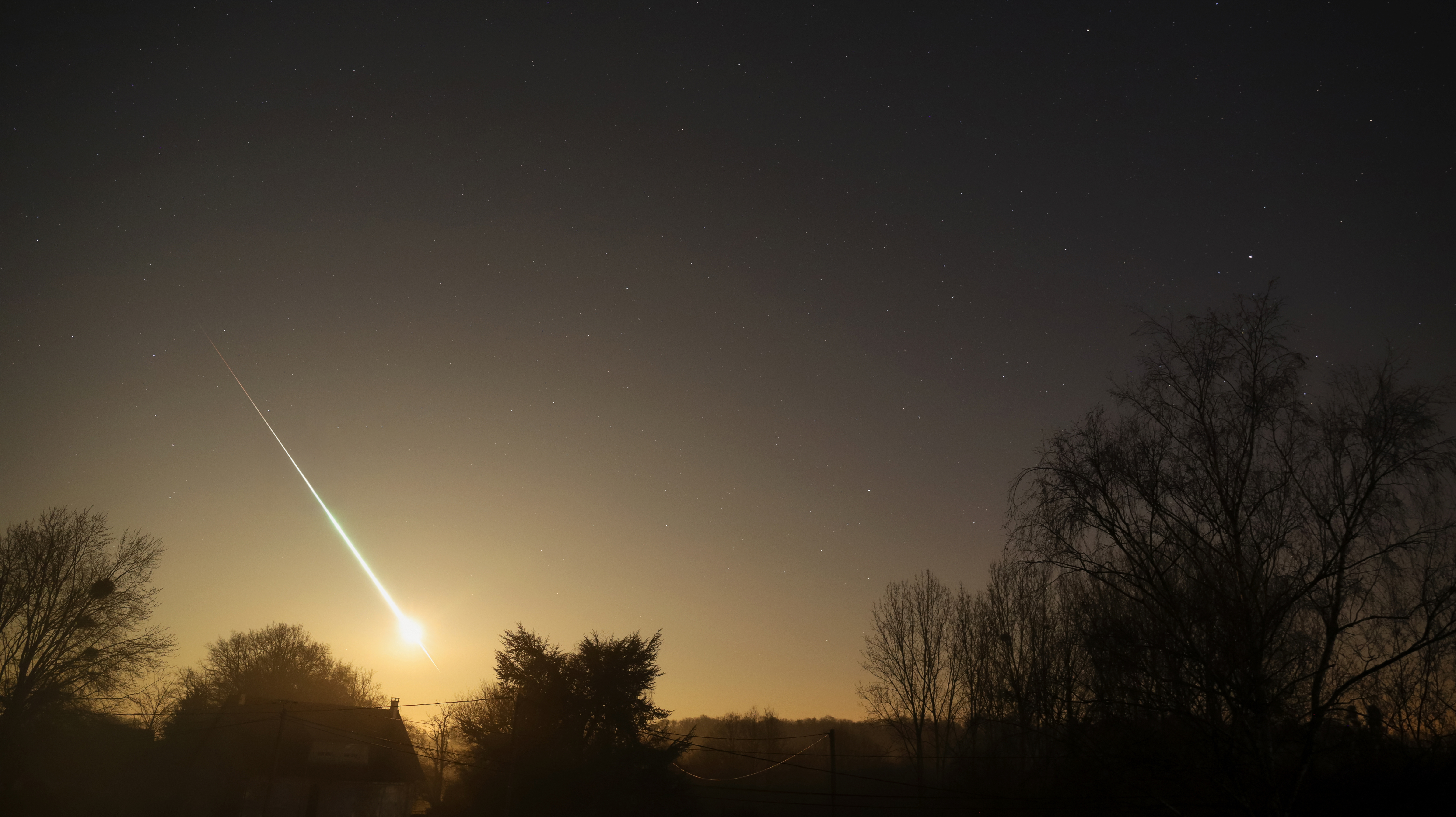 A bright meteor streaks across a dark sky filled with stars, above silhouetted trees and a house.