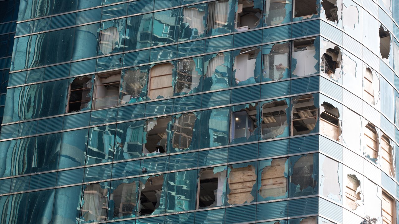 A damaged building facade with broken windows and missing glass panels, reflecting surrounding structures.
