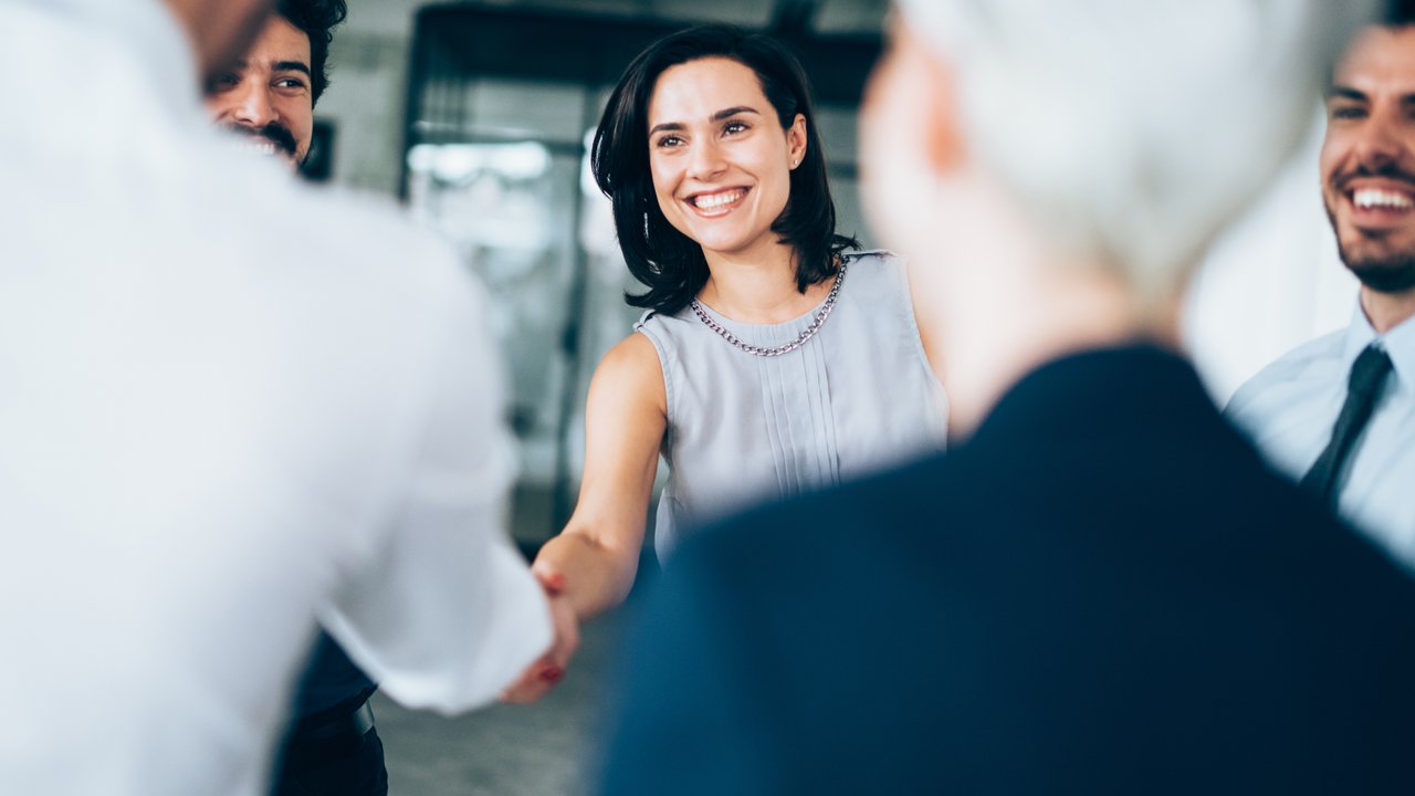 A group of people in business attire, one person shaking hands with another in a bright, modern setting.