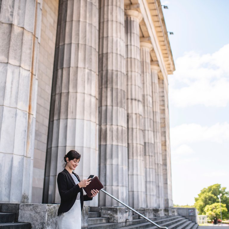 A woman in a black blazer stands on steps, holding a tablet near large stone columns under a blue sky.