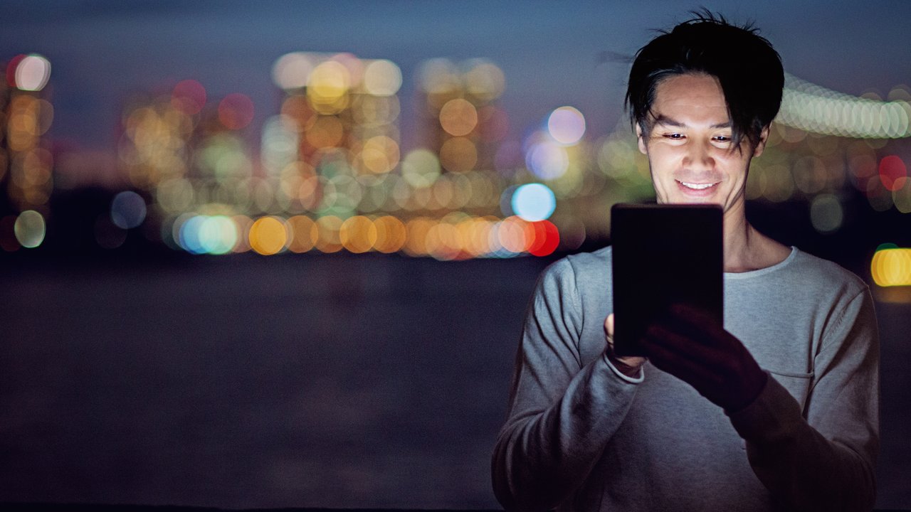 A man smiles while holding a tablet, with a blurred city skyline in the background at night.
