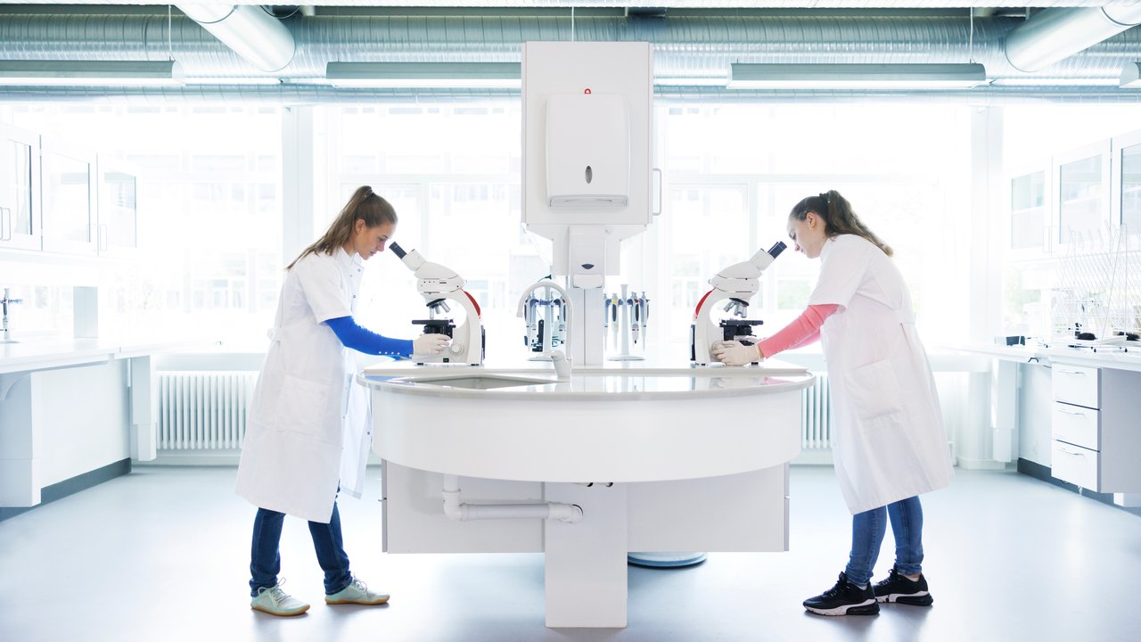 Two women in lab coats using microscopes for genetic multi-cancer early detection at a circular table in a bright laboratory.