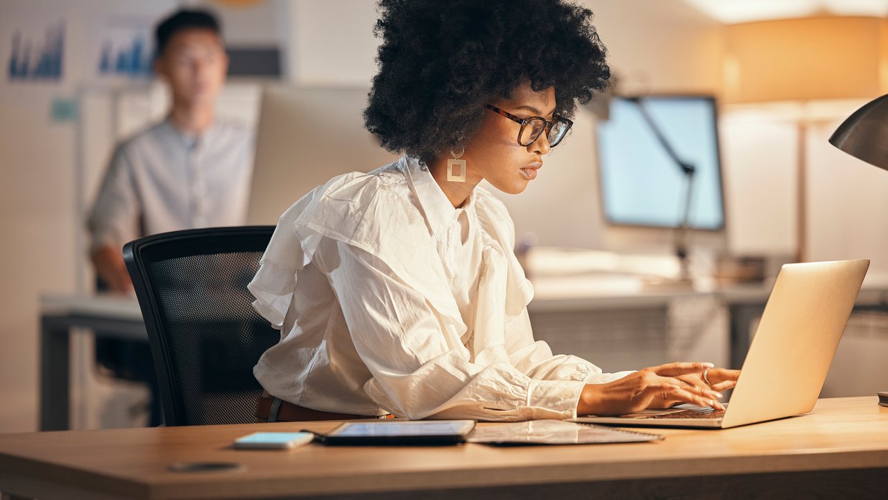 Woman working on a laptop in a modern office A woman with curly hair works on a laptop at a desk in an office, with a man blurred in the background.